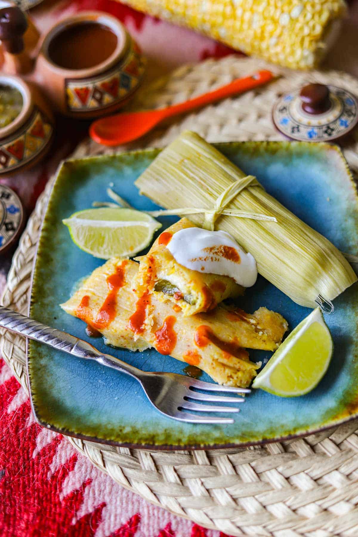corn tamales on a blue plate with lime wedges, salsa, and corn on the cob