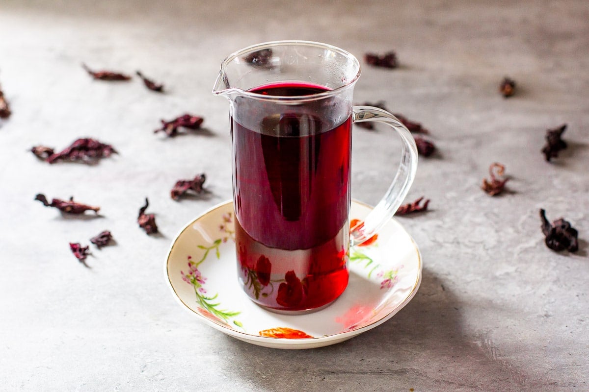 hibiscus syrup in small pitcher beside dried hibiscus flowers.