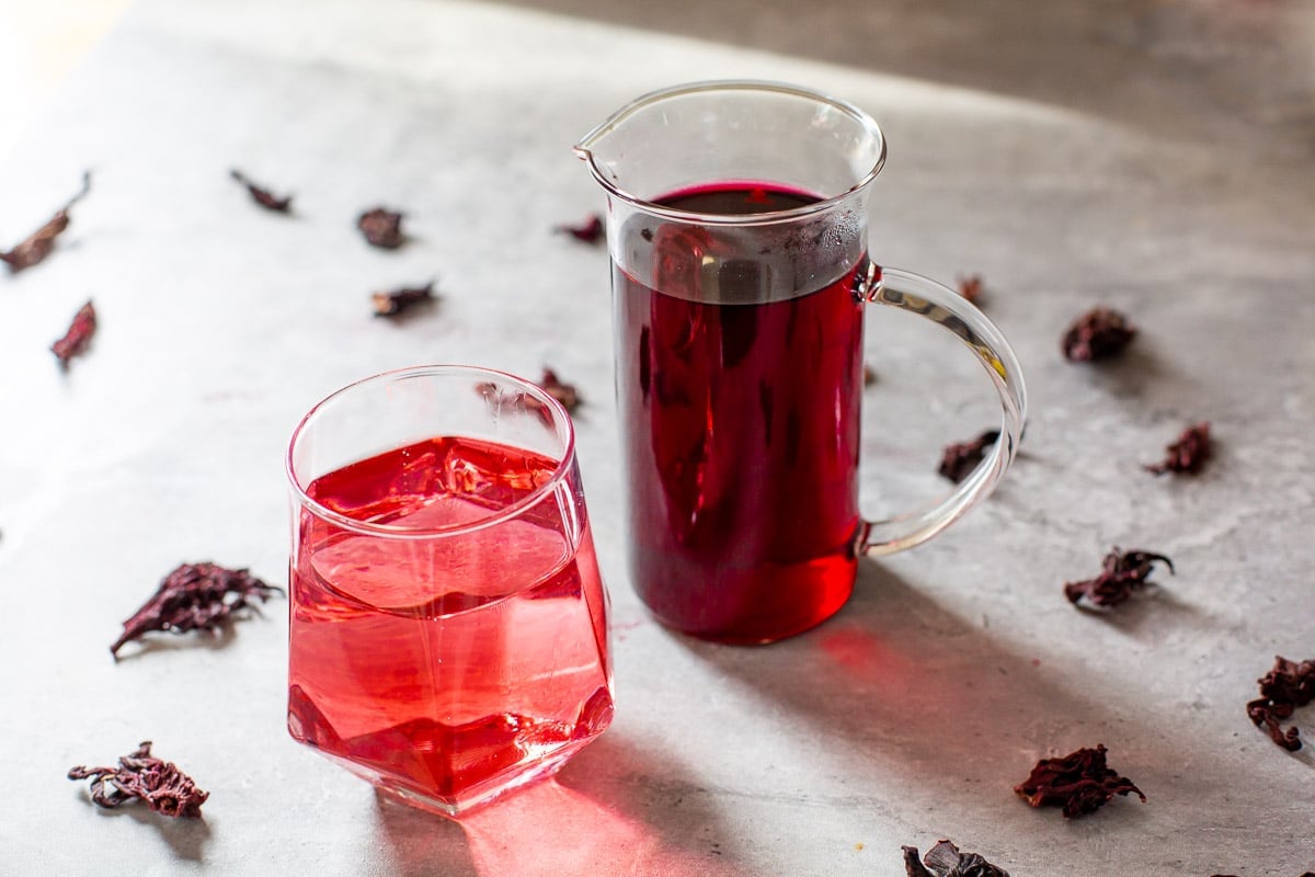 hibiscus syrup in small pitcher beside carbonated drink and dried hibiscus flowers.