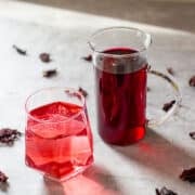 hibiscus syrup in small pitcher beside carbonated drink and dried hibiscus flowers.