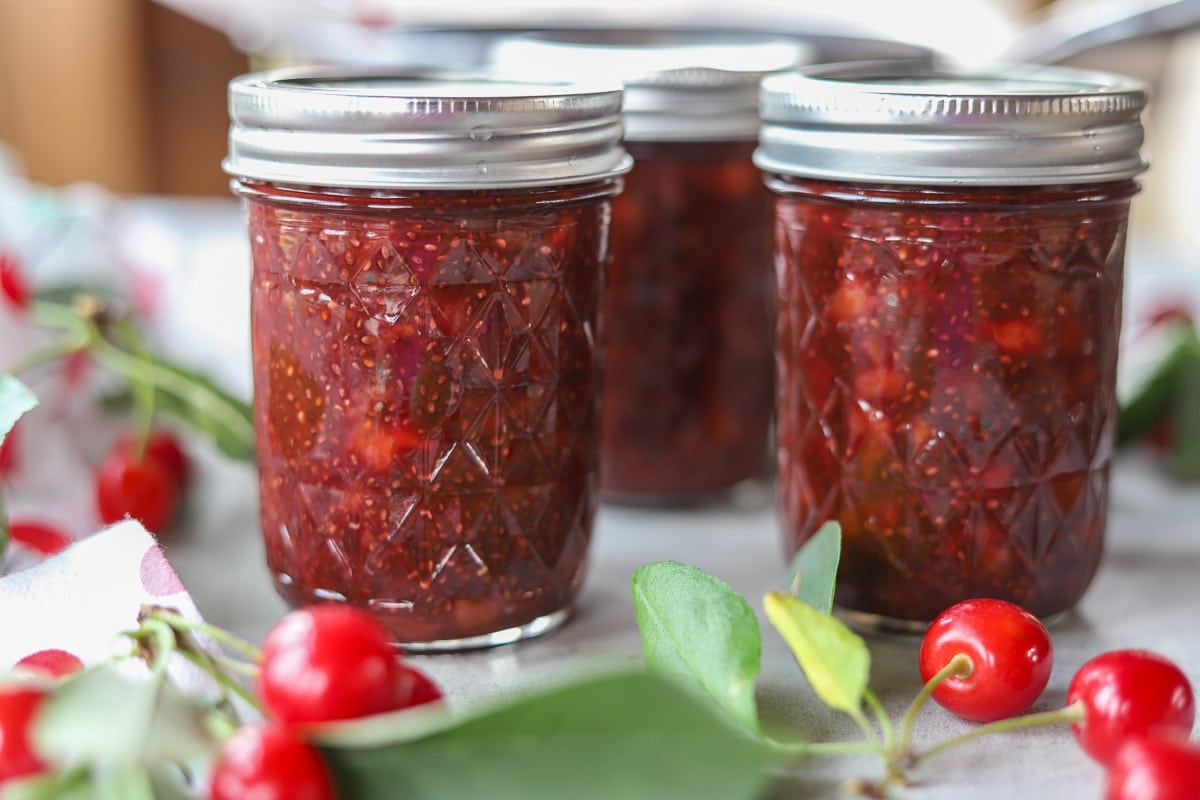 sour cherry jam with chia seeds in 3 mason jars with cherries around it