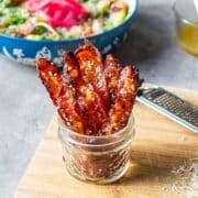 candied bacon strips standing up in jar in front of salad and shredded parmesan cheese.