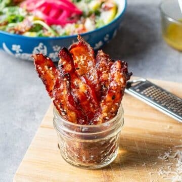 candied bacon strips standing up in jar in front of salad and shredded parmesan cheese.