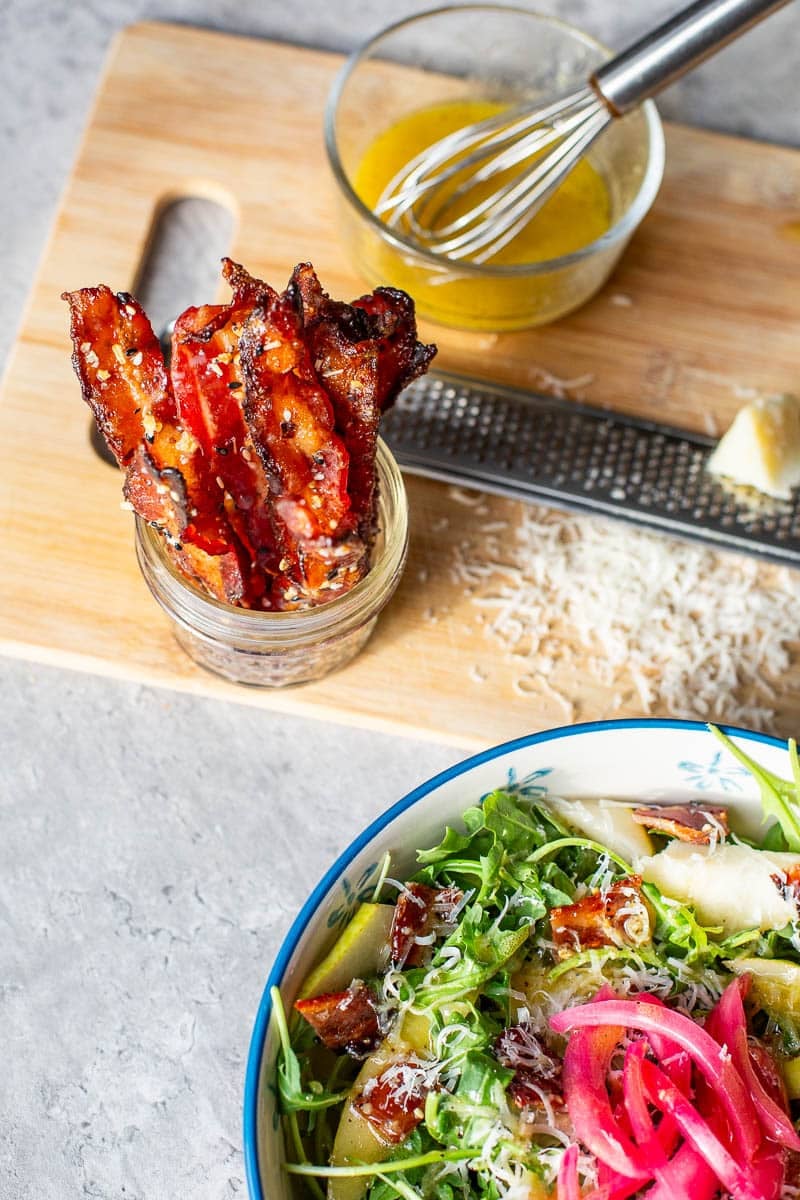 candied bacon strips standing up in jar in front of salad and shredded parmesan cheese.