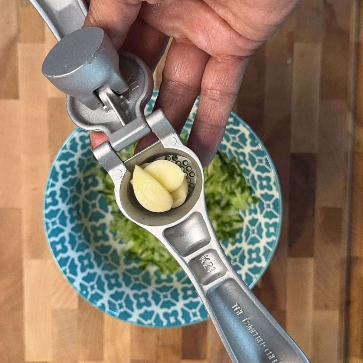 garlic press with 3 cloves of garlic over a blue bowl containing shredded cucumber
