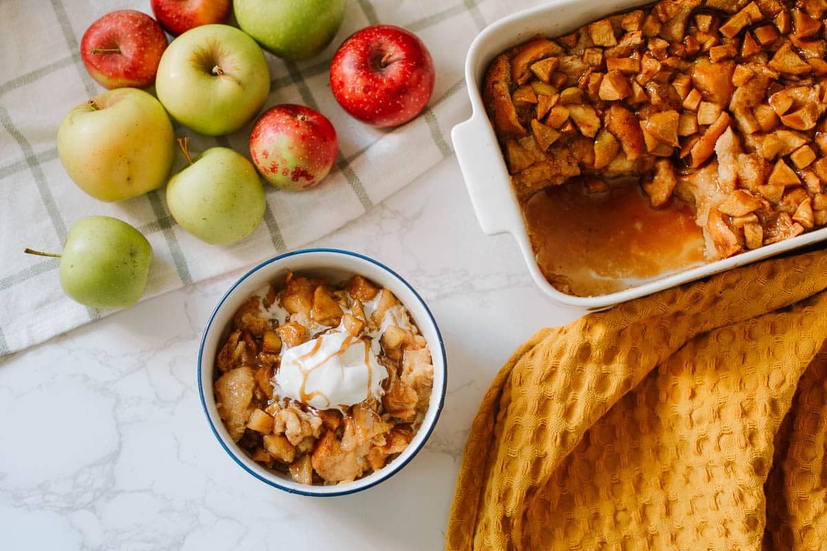 Apple bread pudding with condensed milk in a bowl, with more in a casserole dish and apples on the side.