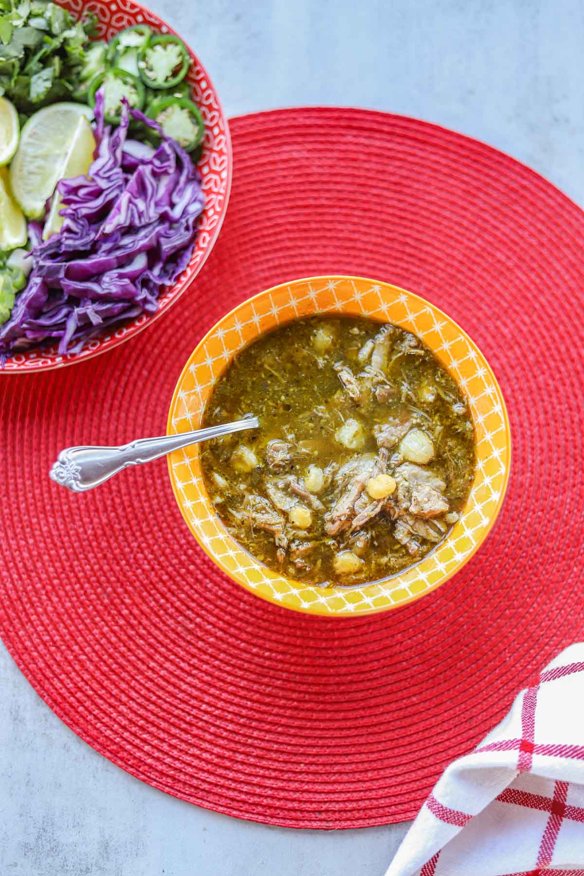 a bowl of pork pozole verde in a yellow bowl on a red mat with sliced veggies next to it
