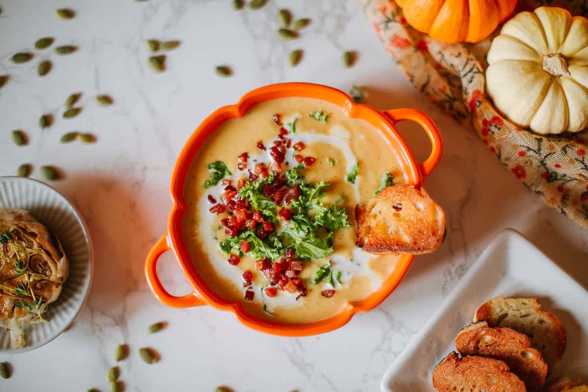 Roasted cauliflower and pumpkin soup in an orange pumpkin-shaped bowl with pumpkin seeds scattered around it and part of a white pumpkin visible.