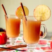 2 glasses of hot spiced apple cider with apples and cinnamon stick next to them