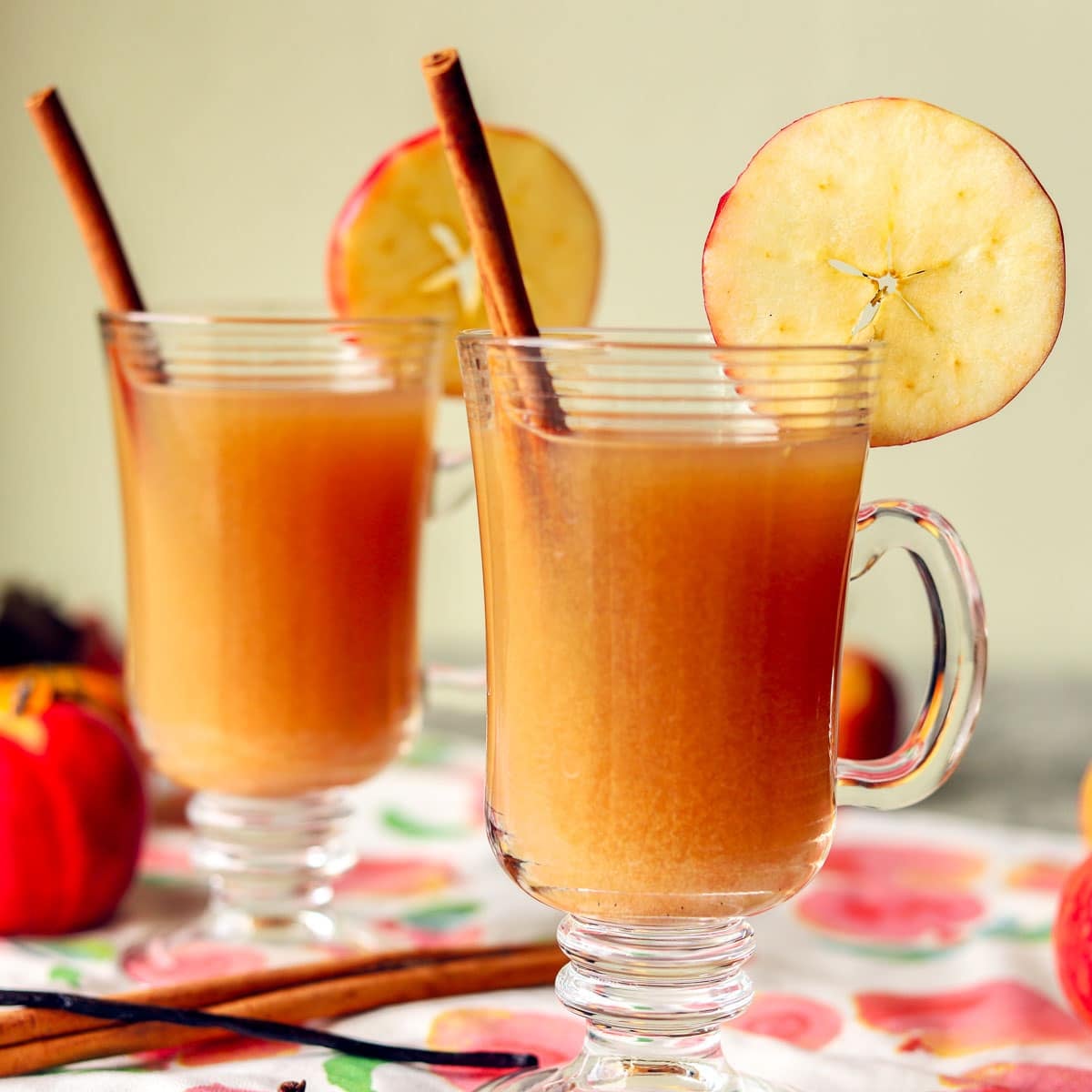 2 glasses of hot spiced apple cider with apples and cinnamon stick next to them