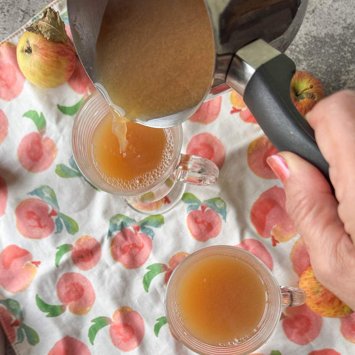 pouring spiced apple cider into 2 mugs with apples in the background