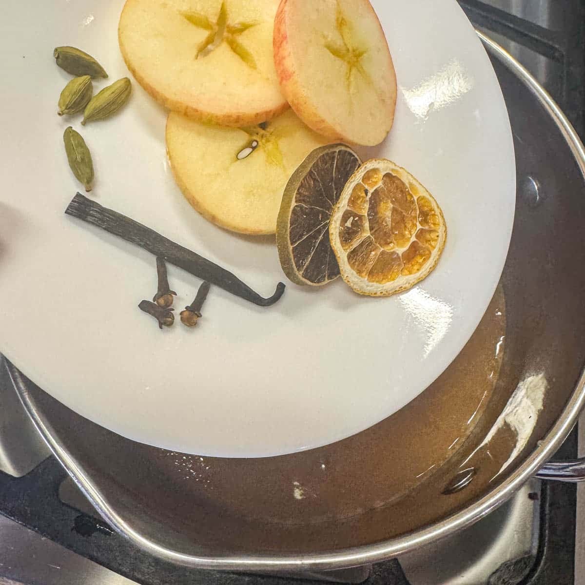 spices, citrus, and apple slices sliding off white plate into a saucepan with apple cider